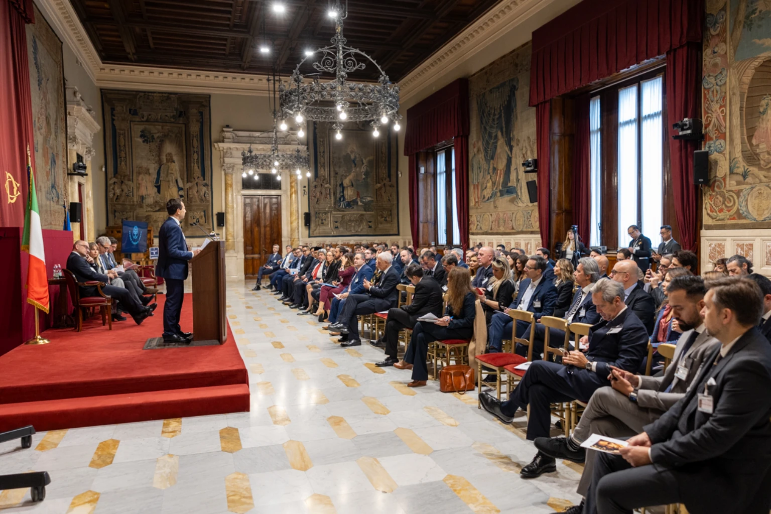 A speaker addresses participants at the Human Economic Forum in an Italian parliamentary hall.